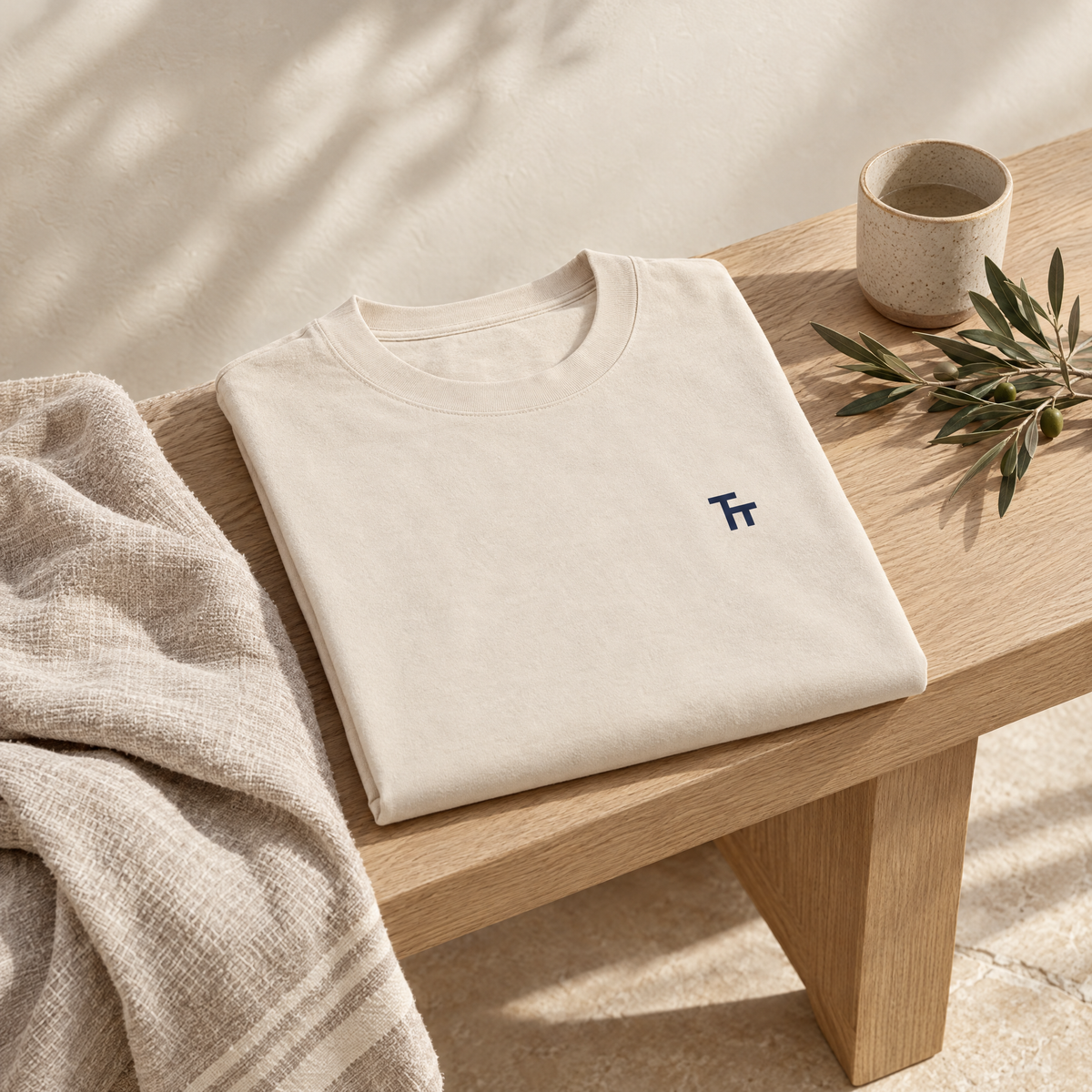 Folded ecru Heavyweight TT Tee on an oak bench with linen and an olive branch